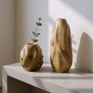 Puede incluir: Dos jarrones de madera con acabado natural. Uno es un jarrón pequeño y redondo con una superficie texturizada, que contiene una ramita de vegetación. El otro es un jarrón alto de forma orgánica. Ambos se exhiben sobre una superficie de hormigón.