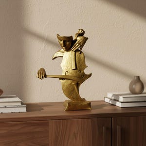Può includere: Una scultura color oro che raffigura una figura umana stilizzata fusa con un violoncello. La figura ha un volto e mani definiti, con una mano che tiene un archetto sul corpo dello strumento. Il pezzo presenta una finitura opaca e testurizzata.