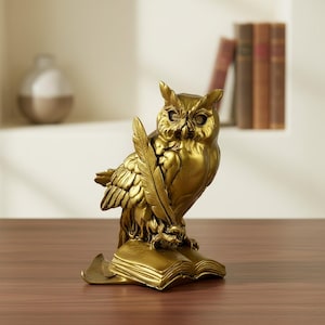 Puede incluir: Una figura de búho de color dorado posada sobre un libro abierto. El búho sostiene una pluma larga y delgada en una de sus garras. La escultura presenta plumas detalladas, ojos definidos y un acabado metálico suave en toda la pieza.