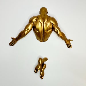 Può includere: Scultura da parete color oro raffigurante un torso umano muscoloso con le braccia tese e un paio di gambe separate. La scultura è montata su una parete bianca, creando un contrasto sorprendente.
