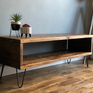 May include: A rustic wooden TV stand with a shelf and hairpin legs. The stand is made of reclaimed wood and has a natural finish. It is sitting on a hardwood floor.
