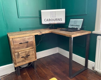 May include: L-shaped wooden desk with a rustic, reclaimed wood appearance. The desk features a two-drawer cabinet on the left and a metal frame. A computer monitor and laptop with the text "CABOURNS" are on the desk.