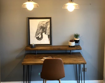 May include: A rustic wooden desk with black hairpin legs and a top shelf. The desk has a brown leather chair and a framed print of an elephant on the wall behind it.
