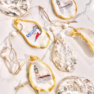May include: Hand-painted oyster shell ornaments with gold trim and twine hangers. Some shells feature lighthouse illustrations, while another displays "LUNCH LOBSTER ROLL" text. The shells are white and beige with a natural texture.