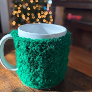 May include: A white ceramic mug with a green crocheted cosy. The mug is sitting on a wooden table.