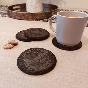 May include: Three circular black slate coasters featuring engraved military aircraft designs. One coaster displays a Typhoon jet with the text HANGAR979, AGILE, LETHAL, and VERSATILE. Another shows a Thunderbolt II. A white ceramic mug sits on one coaster.