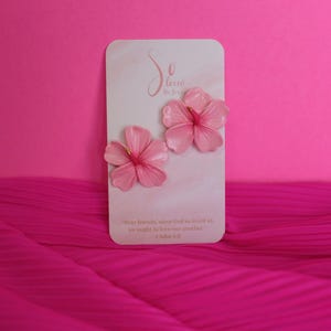 May include: A pair of pink hibiscus flower earrings with gold-colored centers rest on a white card. The card has the text "So loved by Julie" and a Bible verse. The background is a vibrant pink.