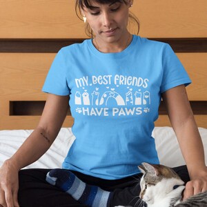 May include: A woman wearing a light blue t-shirt with the text "My best friends have paws" and a graphic of paw prints. She is sitting on a bed with a cat.