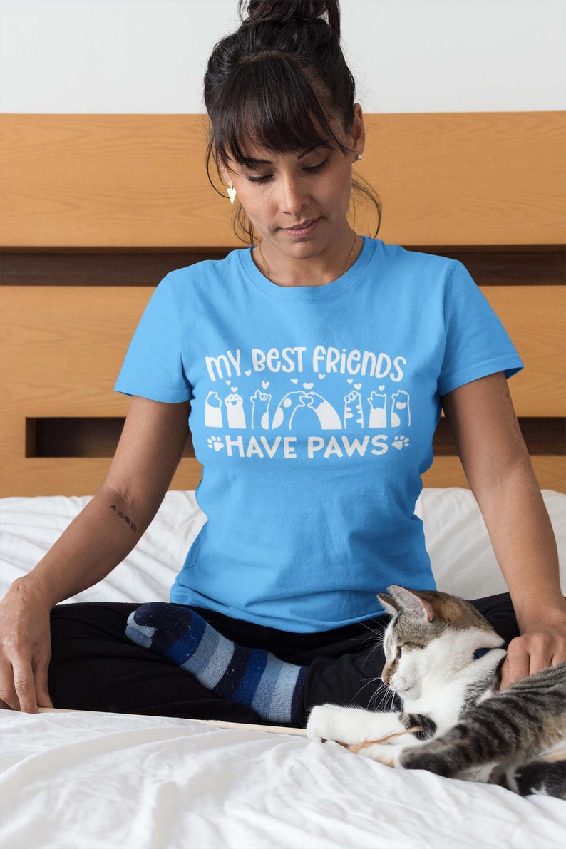 May include: A woman wearing a light blue t-shirt with the text "My best friends have paws" and a graphic of paw prints. She is sitting on a bed with a cat.