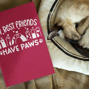 May include: A red journal with the text "My best friends have paws" in white lettering. The text is surrounded by paw prints and hearts. A cat is sleeping on a cushion in the background.
