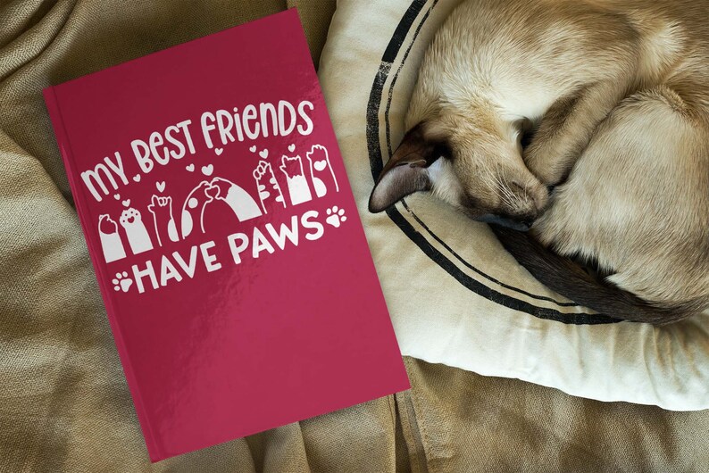 May include: A red journal with the text "My best friends have paws" in white lettering. The text is surrounded by paw prints and hearts. A cat is sleeping on a cushion in the background.