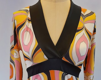May include: A colorful, patterned dress with a V-neck and three-quarter sleeves. The dress features a black trim at the neckline and waist, and a vibrant print in shades of orange, yellow, pink, and white. The dress is displayed on a mannequin.