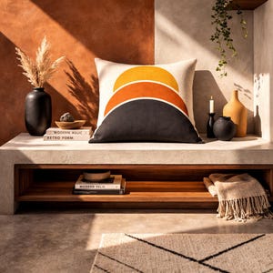 Könnte beinhalten: Ein dekoratives Kissen mit einem geometrischen Design aus drei übereinander angeordneten, halbkreisförmigen Formen in Gelb, Orange und Schwarz. Das Kissen liegt auf einem Betonregal mit einer schwarzen Vase, Büchern und einer Kerze. Die Wörter "MODERN RELIC" und "RETRO FORM" sind auf den Büchern zu sehen.