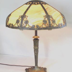 May include: A table lamp featuring a wide, dome-shaped shade with amber-colored glass panels and dark metal overlays. The metal base is fluted and ornate, with a square, scalloped foot. The lamp includes a dark brown power cord extending from the base.