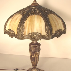 May include: An antique table lamp with a domed shade featuring a stained glass design in cream and tan hues. The lamp has a bronze-colored metal base and intricate floral and leaf detailing. The lamp is plugged into an electrical outlet.