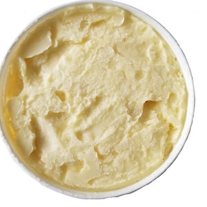 May include: A close-up view of a white container filled with a light yellow, creamy substance. The texture appears thick and whipped, with visible ridges and peaks. The container is round and the background is white.