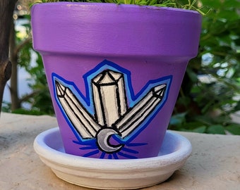 May include: Purple terracotta planter with a white and blue painted design of crystals and a crescent moon. The planter is filled with green air plants.