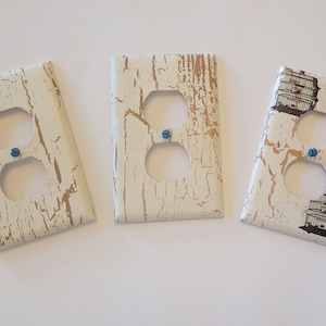 Può includere: Tre placche per prese di corrente bianche con un design a venature di legno invecchiato. La placca centrale ha una finitura metallica dorata. La placca a destra presenta un'immagine in bianco e nero di una gabbia per uccelli.