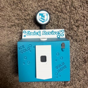 May include: A blue vending machine replica featuring the text Quick Revive! at the top, with the phrases ICE COLD and SOLD HERE embossed on the front. It includes a white central panel, a small black switch, and a circular sign with a white icon.