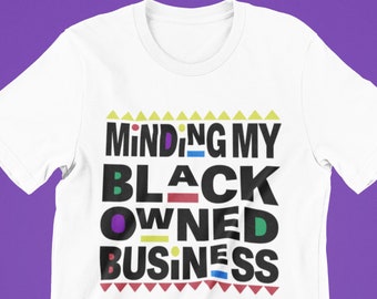 May include: White t-shirt with the text "Minding My Black Owned Business" in colourful block letters with a geometric pattern above and below the text.
