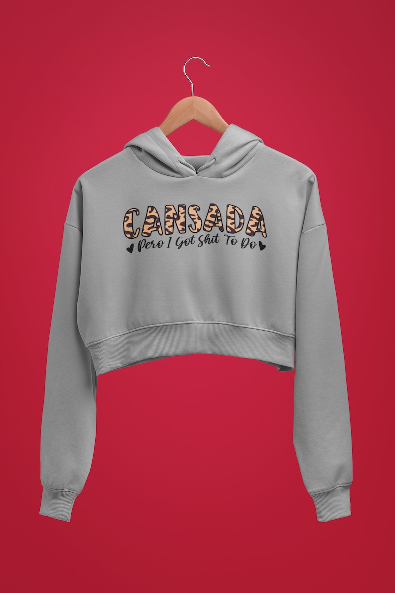 May include: A gray cropped hoodie with a leopard print design that says "Cansada pero I got shit to do" in black text.