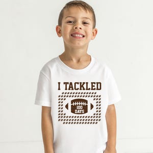 A young boy wearing a white t-shirt with a graphic design that says "I TACKLED 100 DAYS" and features a football.