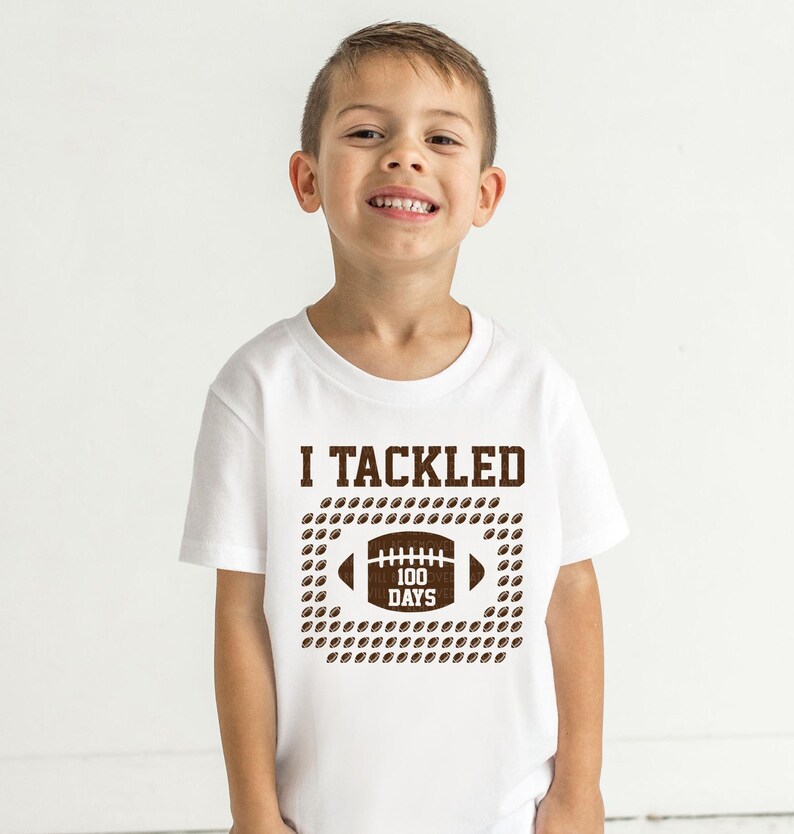 A young boy wearing a white t-shirt with a graphic design that says "I TACKLED 100 DAYS" and features a football.