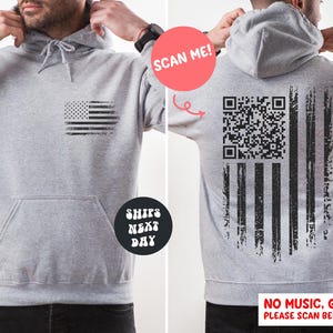 May include: A gray hoodie with a distressed American flag design on the front and back. The back features a QR code within the flag's stripes. The hoodie has a front pocket and a drawstring hood. The text "SCAN ME!" is visible.