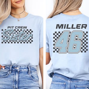 A woman wearing a light blue t-shirt with a checkered pattern and the text "PIT CIRCLE Sister" printed on it.