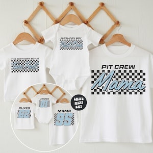 A collection of white t-shirts with various text and numbers printed on them, including a "PIT CREW Mama" t-shirt and a "The birthday boy is Tho fast" t-shirt. The t-shirts are displayed on wooden hangers against a white background.