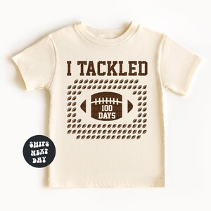 A beige t-shirt with a graphic design featuring a football and the text "I TACKLED 100 DAYS" printed on it.