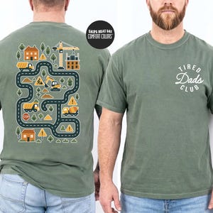 May include: Olive green t-shirt with a construction-themed graphic on the back, featuring roads, buildings, and construction vehicles. The front of the shirt displays the text "Tired Dads Club" in a white, cursive font. The shirt is made of a soft, comfortable material.