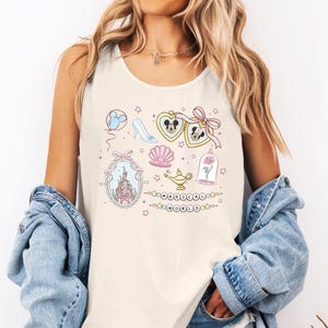 A woman wearing a white tank top with a Disney-themed design, including a castle, Mickey Mouse, and other whimsical elements. She is also wearing a denim jacket and has her hands in her pockets.