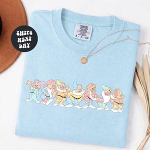 May include: Light blue Comfort Colors t-shirt featuring a line of seven cartoon dwarfs in various colors. The shirt has a small tag on the upper right and a gold necklace. The words "Ships Next Day" are in a black circle.