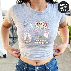 A woman wearing a gray t-shirt with a graphic design featuring various Disney-themed elements such as Mickey Mouse, a castle, and a rose.