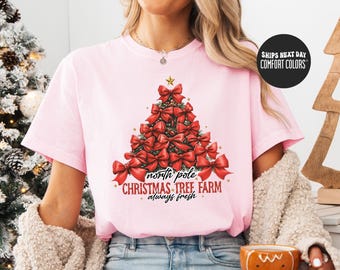 May include: A light pink t-shirt featuring a Christmas tree design made of red bows. The text on the shirt reads "north pole CHRISTMAS TREE FARM always fresh". The shirt is a Comfort Colors brand.