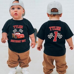 A young child wearing a black t-shirt with a red and white Cars-themed design and the name " Tyler" printed on it, along with the number "1" and the word " birthday boy" below. The child is also wearing a gray baseball cap.