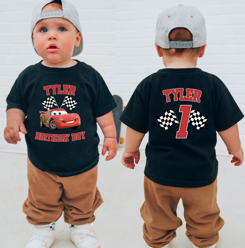A young child wearing a black t-shirt with a red and white Cars-themed design and the name " Tyler" printed on it, along with the number "1" and the word " birthday boy" below. The child is also wearing a gray baseball cap.