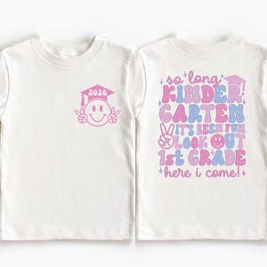 A white t-shirt with a graduation cap and a smiley face on the left side, and text on the back that says "so long Kindergarten Garten, it's been fun 1st Grade here I come!"