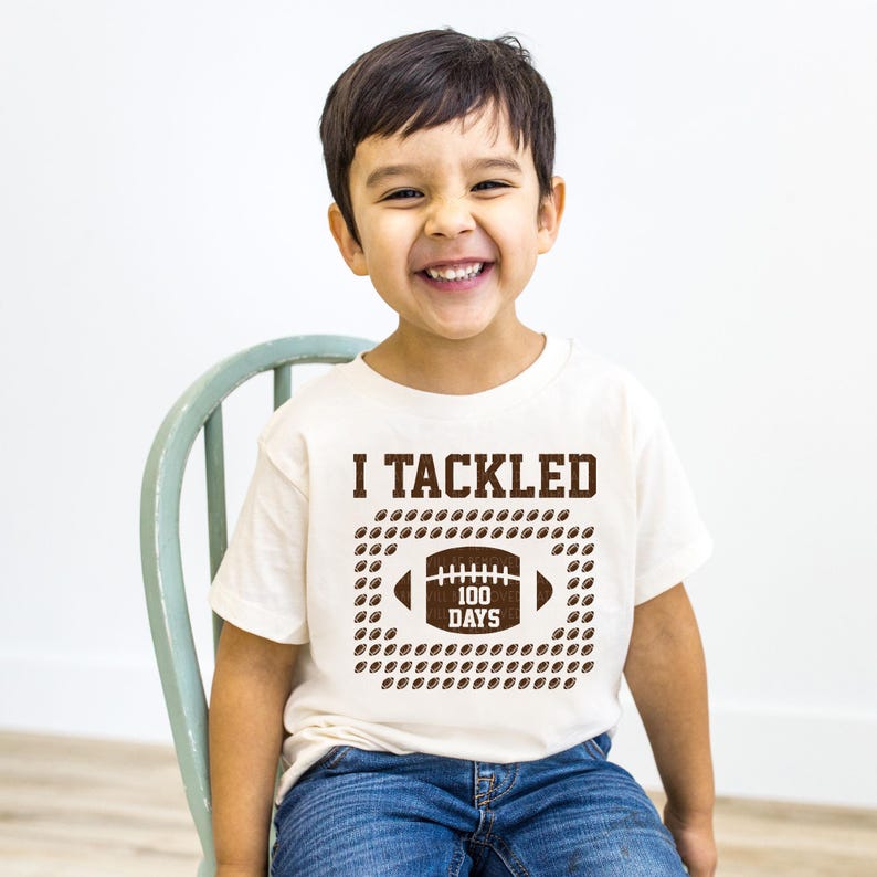 A young boy is sitting on a chair, smiling and wearing a white t-shirt with the text "I TACKLED 100 DAYS" printed on it.