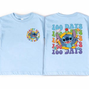 A light blue t-shirt with a colorful graphic design featuring a cartoon character and the text "100 DAYS" in a playful, celebratory style.