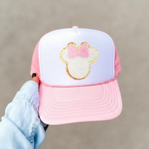May include: Pink and white trucker hat featuring a Minnie Mouse design. The hat has a pink brim and mesh back, with a white front panel. The Minnie Mouse design is outlined in gold glitter and features a pink bow.