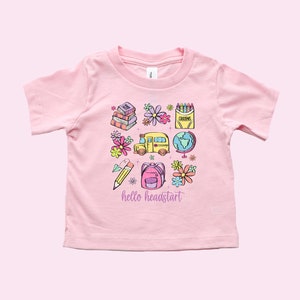 May include: Pink short-sleeved t-shirt with a graphic design of school supplies, including a school bus, books, a pencil, a backpack, crayons, a globe, and flowers. The text "hello headstart" is printed on the shirt.