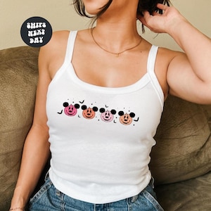 May include: A white ribbed tank top featuring a graphic print of four stylized pumpkins with mouse-ear silhouettes in shades of pink and orange, accented by small black bats and stars. The design is centered on the chest of the garment.