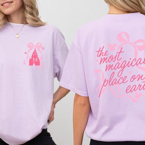 A woman wearing a pink t-shirt with a castle and bow design on the front and text on the back.