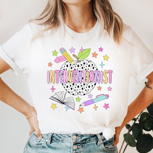 May include: White t-shirt with a black and white polka dot apple, a pencil, a pen, a book, and stars. The text "INTERVENTIONIST" is written in rainbow colors on the apple.