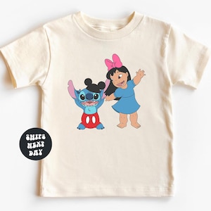 A white t-shirt with a graphic design featuring two cartoon characters, a girl and a blue character, along with a text overlay that reads "SHIPS NEXT DAY".