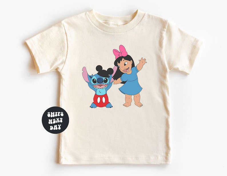 A white t-shirt with a graphic design featuring two cartoon characters, a girl and a blue character, along with a text overlay that reads "SHIPS NEXT DAY".