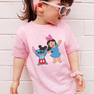 A young girl wearing a pink t-shirt with a cartoon character printed on it, standing against a white brick wall.