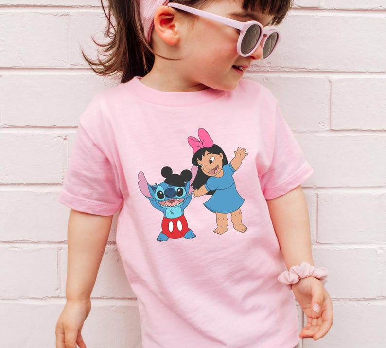 A young girl wearing a pink t-shirt with a cartoon character printed on it, standing against a white brick wall.